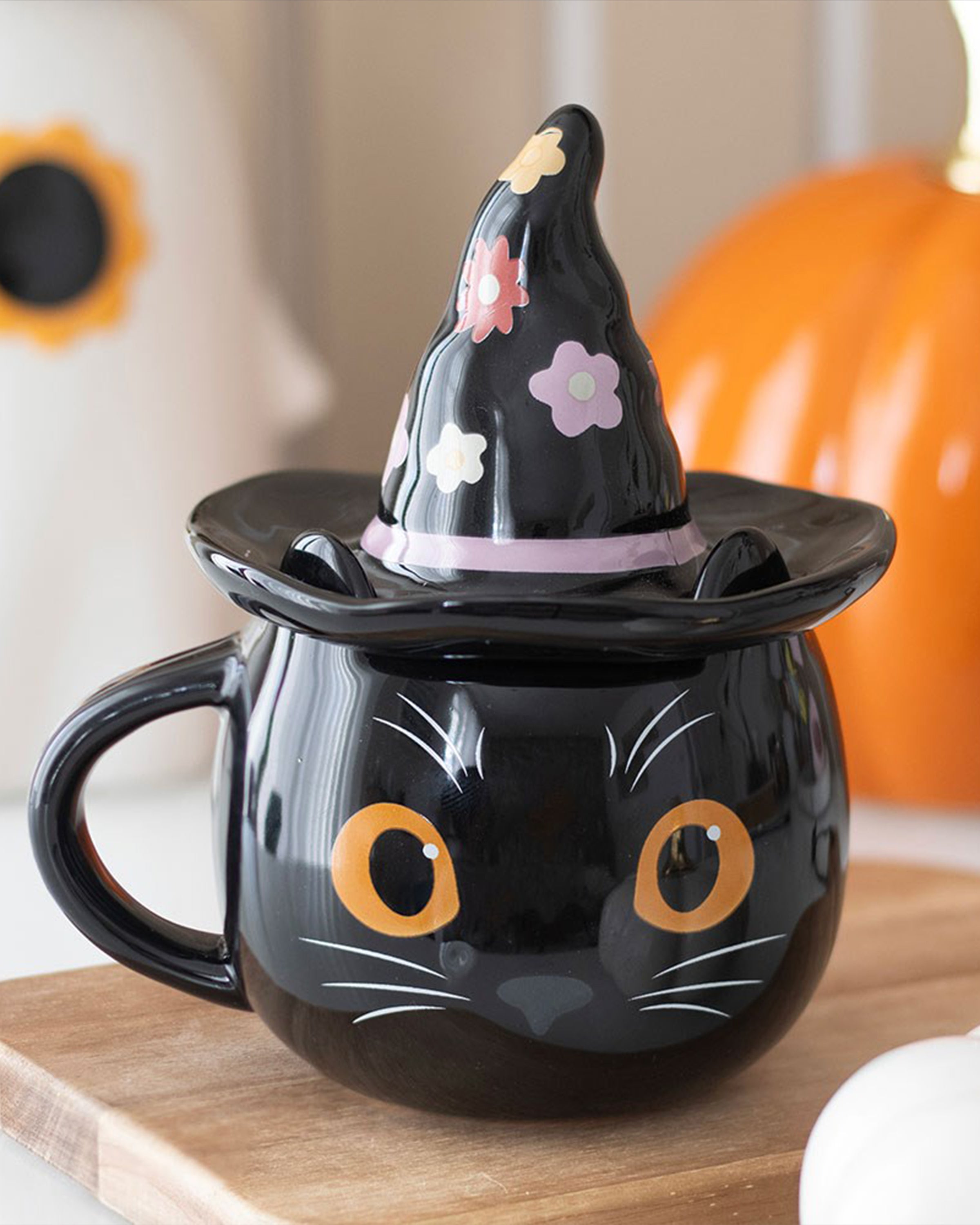 Styled photo of the Groovy Witch Cat Lidded Mug by Rebel Romance placed on a wooden board with pumpkins in the background. This whimsical Halloween-themed mug features a black cat face and floral witch hat lid – ideal for autumn aesthetics.