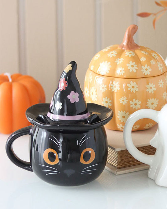 Close-up of the Rebel Romance Groovy Witch Cat Lidded Mug styled on a white table with pumpkins and books. The mug is shaped like a black cat with orange eyes and wears a black floral witch hat as a lid. A charming and spooky autumn-themed homeware gift idea from a small UK business.