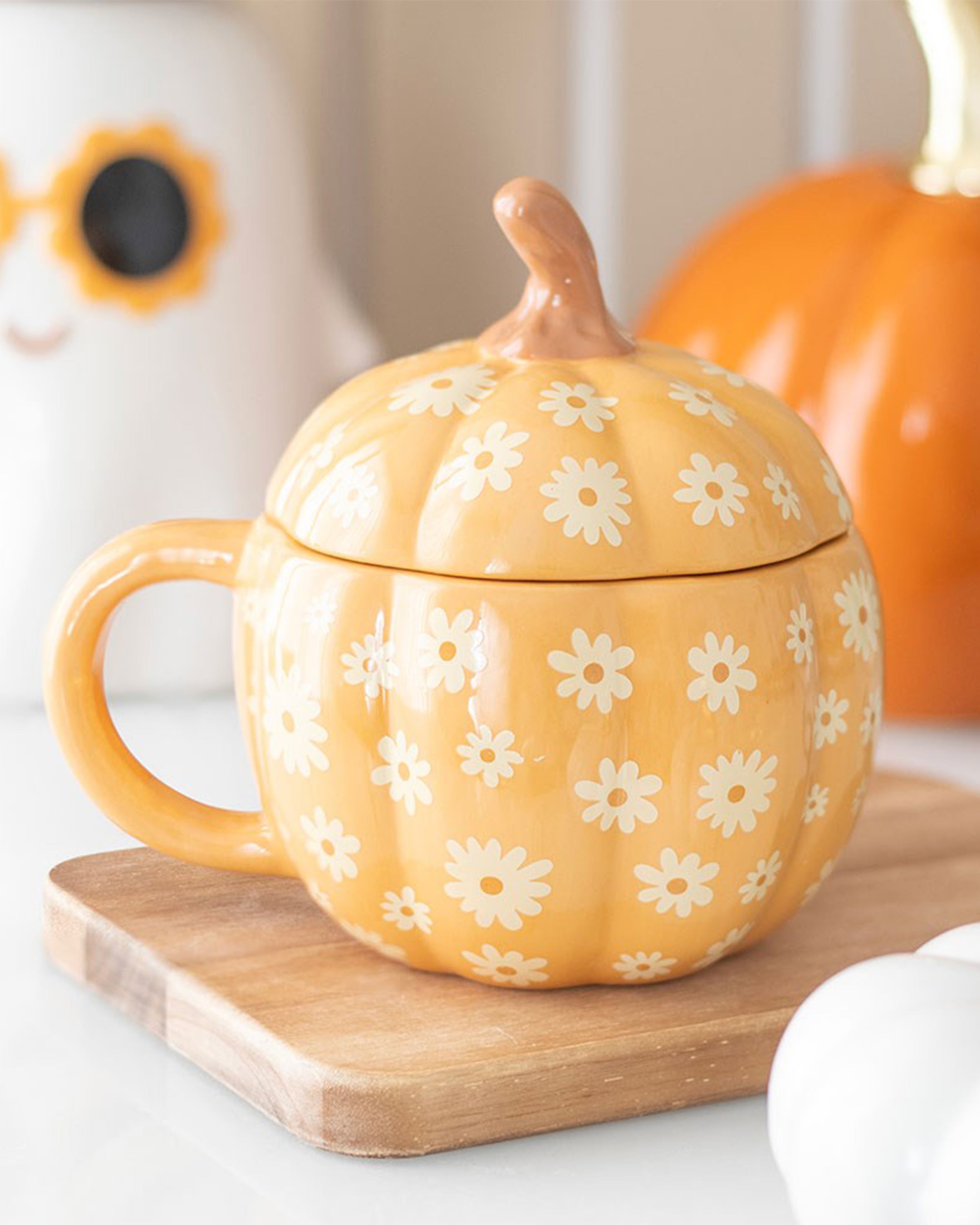 A lifestyle image of the Rebel Romance Groovy Floral Pumpkin Mug styled on a wooden board with autumn decorations. This quirky ceramic mug features a pumpkin shape, floral print, and a fun lid that keeps drinks warm in seasonal style.