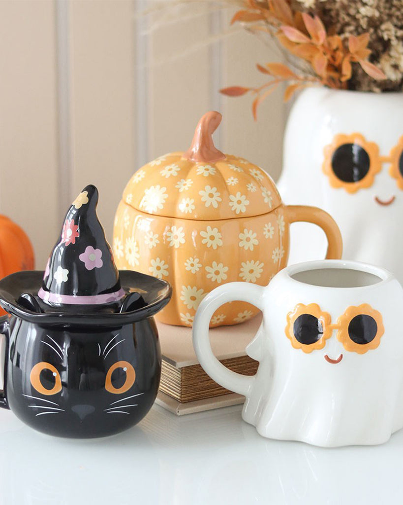 A styled photo featuring Rebel Romance’s Groovy Floral Pumpkin Lidded Mug alongside other quirky Halloween-themed mugs, including a black cat witch mug and a ghost mug. The pumpkin mug is orange with cream flower patterns and a curved lid shaped like a stem. Fun, retro, and perfect for autumn.