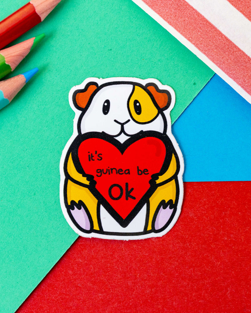 Cute vinyl sticker featuring a cartoon guinea pig holding a red heart that says “It’s guinea be OK”, designed by Innabox and sold by Rebel Romance. This cheerful waterproof sticker is shaped like a guinea pig and is perfect for laptops, water bottles, or notebooks. Bright and uplifting design to add a smile to your day. Ideal for fans of alternative stationery and small UK brands.