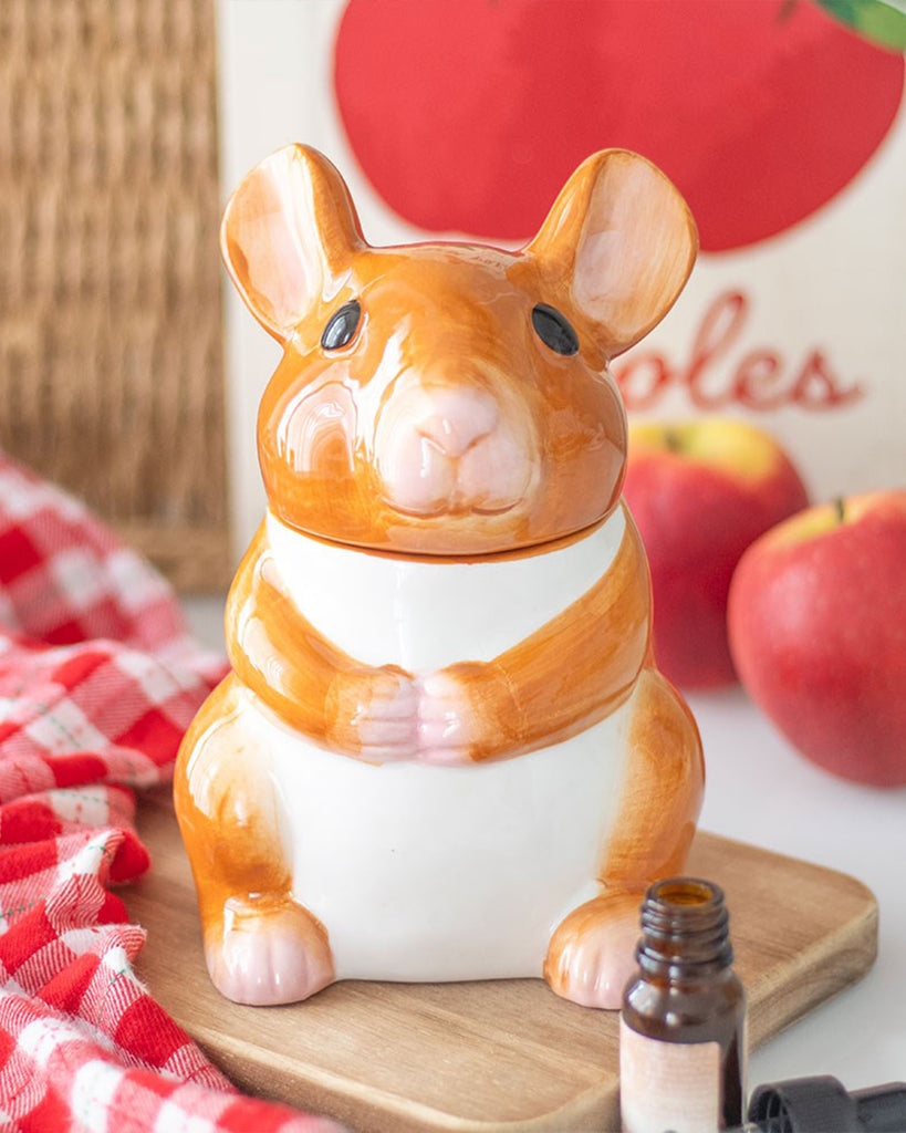 Mouse-shaped ceramic oil burner in orange and white, featuring a cute face and glossy finish, shown on a kitchen counter with apples and a red gingham cloth.