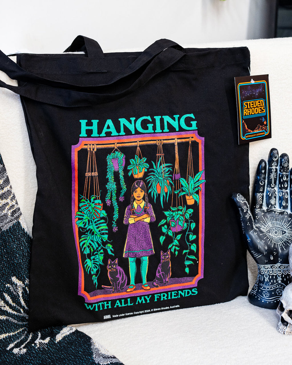 A black tote bag featuring Steven Rhodes’ “Hanging With All My Friends” design. The artwork shows a young girl in a purple dress and green tights standing with two black cats amidst lush hanging plants and greenery. The text reads “Hanging With All My Friends” in bright green lettering.