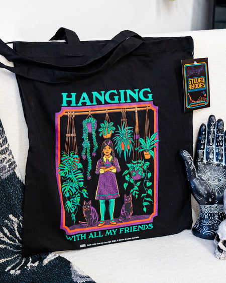 A black tote bag featuring Steven Rhodes’ “Hanging With All My Friends” design. The artwork shows a young girl in a purple dress and green tights standing with two black cats amidst lush hanging plants and greenery. The text reads “Hanging With All My Friends” in bright green lettering.