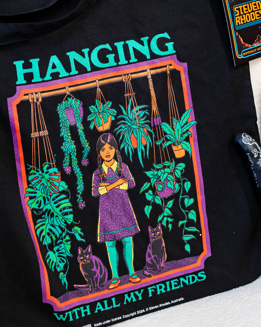 A black tote bag featuring Steven Rhodes’ “Hanging With All My Friends” design. The artwork shows a young girl in a purple dress and green tights standing with two black cats amidst lush hanging plants and greenery. The text reads “Hanging With All My Friends” in bright green lettering.