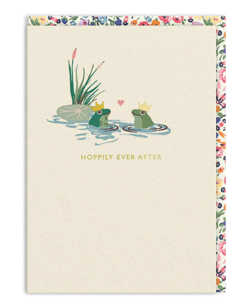 Cath Kidston Hoppily Ever After greeting card from Rebel Romance, featuring two cartoon frogs wearing gold crowns in a lily pond with a heart between them. Surrounded by reeds and a floral border, this whimsical A6 card is perfect for weddings, engagements, or romantic fairytales. Blank inside with a floral envelope. Ideal for quirky, alternative love stories.