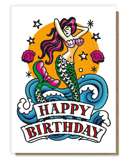 A vibrant greeting card featuring a retro tattoo-style mermaid with flowing purple hair, shimmering green scales, and a pink seashell top. The mermaid is surrounded by ocean waves, seashells, stars, and an orange sunburst background. The banner below reads “Happy Birthday” in bold red and black vintage typography, paired with a recycled kraft envelope.