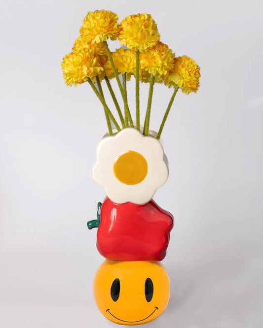 Rebel Romance Smiley Fun Vase filled with yellow faux flowers. Playful ceramic vase shaped like a stack of a smiley face, apple, and daisy. A cheerful and quirky centrepiece perfect for dopamine décor lovers and alternative home aesthetics.