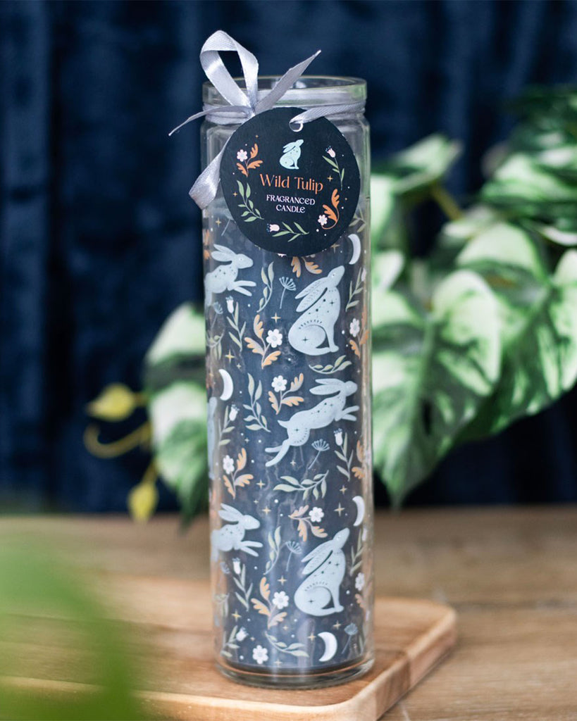 Styled shot of the Midnight Hare Wild Tulip Tube Candle by Rebel Romance, placed on a wooden surface with leafy plants. The mystical hare design and silver ribbon make it a perfect alternative gift or home decor piece.