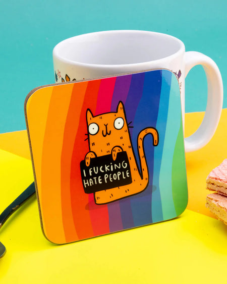 Brightly coloured coaster featuring Katie Abey’s orange cartoon cat holding a black sign that says “I F*cking Hate People,” set against a vertical rainbow-striped background. The coaster is placed in front of a white mug and yellow backdrop.