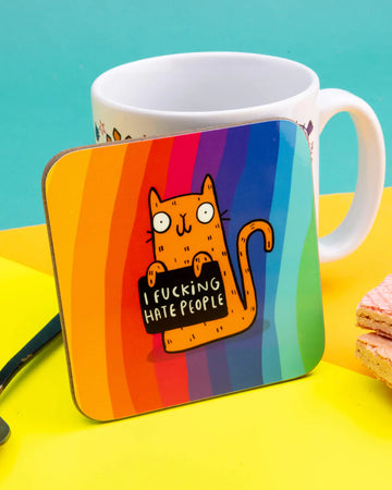 Brightly coloured coaster featuring Katie Abey’s orange cartoon cat holding a black sign that says “I F*cking Hate People,” set against a vertical rainbow-striped background. The coaster is placed in front of a white mug and yellow backdrop.