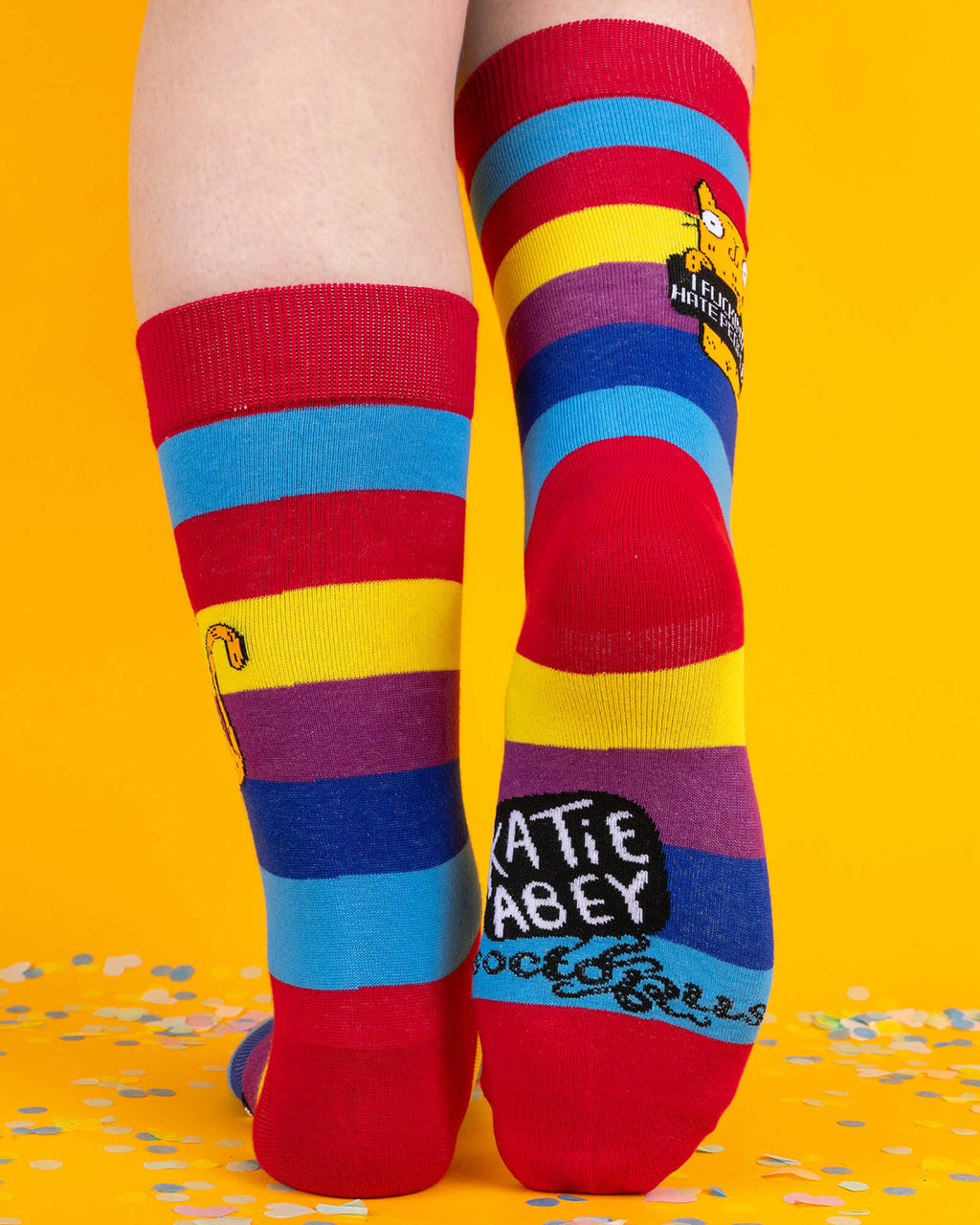Back view of rainbow Katie Abey socks showing the same orange cat and the words “Katie Abey” on the sole. The vibrant stripes pop against a yellow background and scattered confetti.