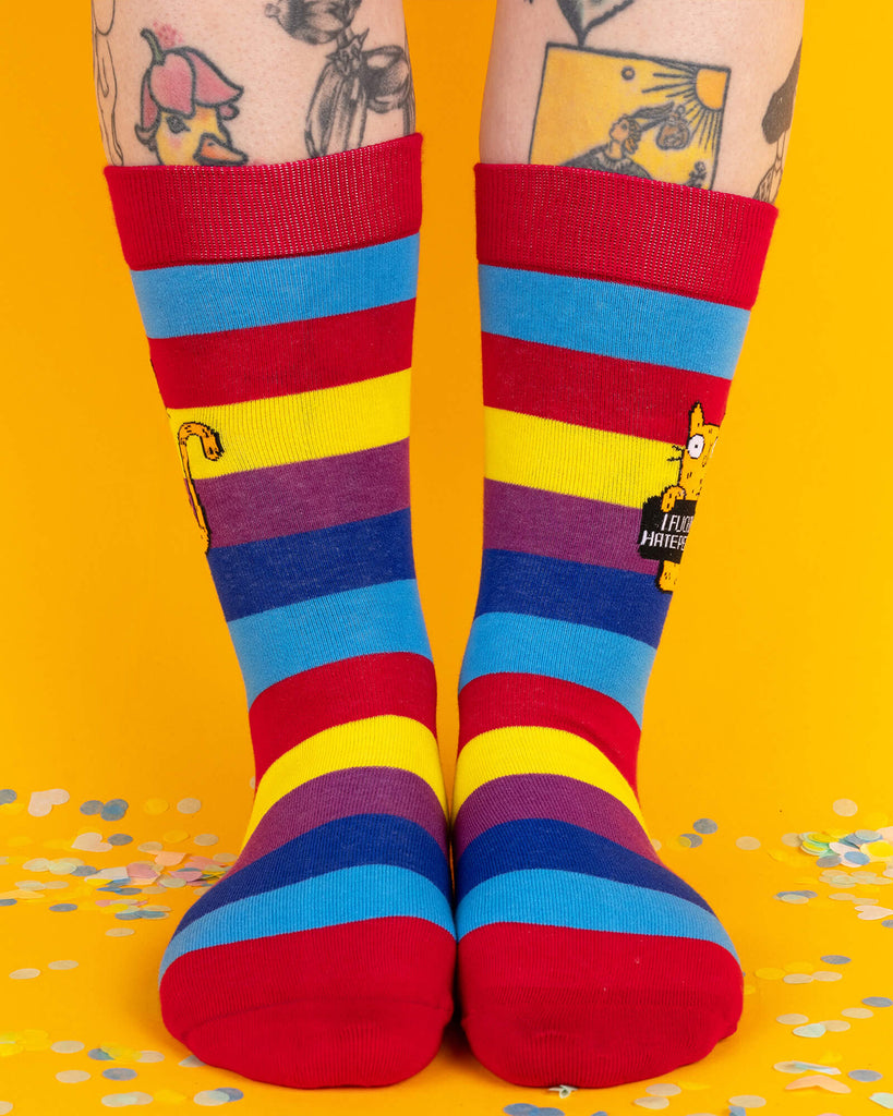 A pair of colourful rainbow-striped socks by Katie Abey worn on tattooed legs. The design features bold red, yellow, blue, and purple stripes, with an orange cat holding a sign that reads “I F*cking Hate People.” Fun and bold footwear for alternative fashion lovers.