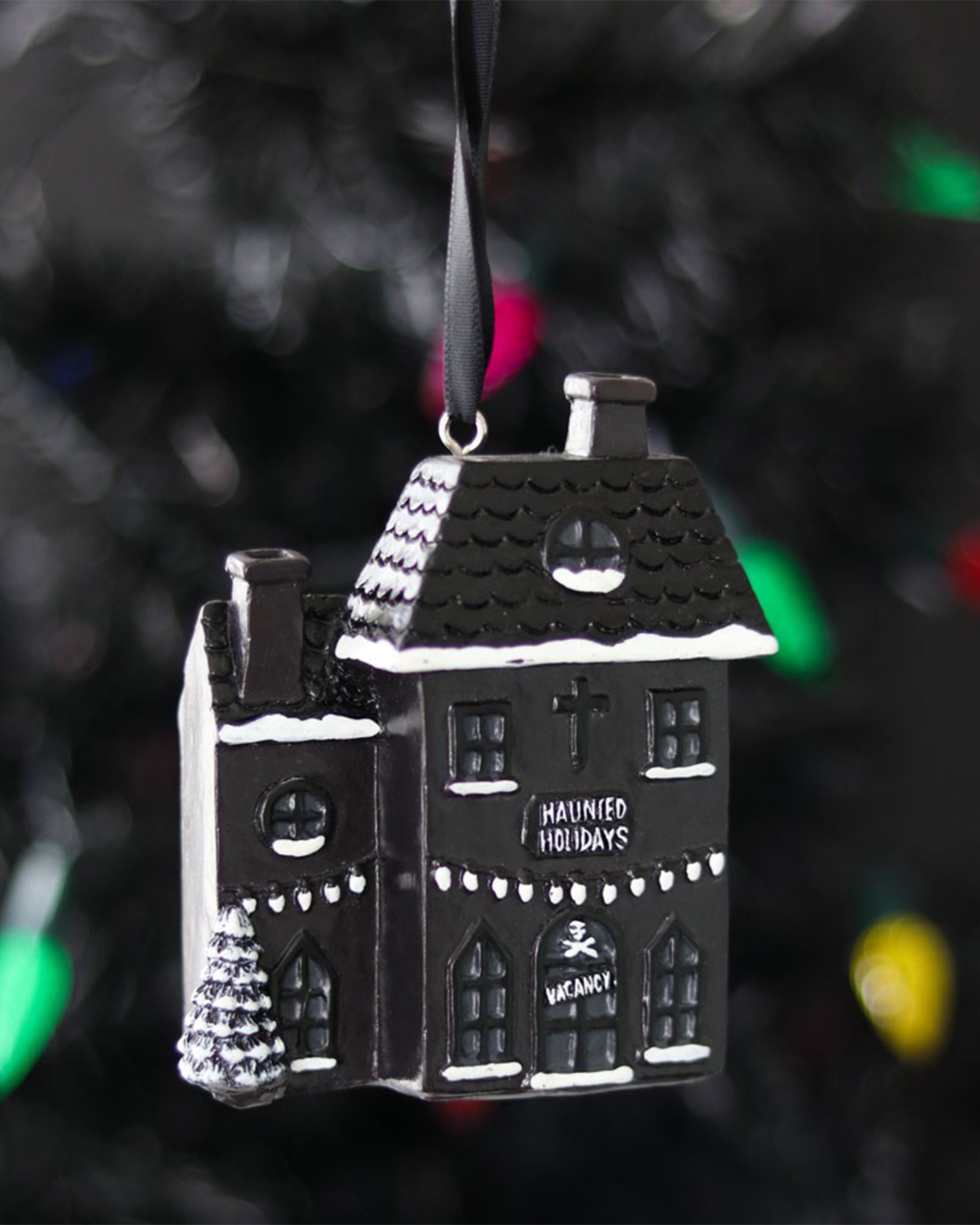 Close-up of the Rebel Romance Haunted Holiday House resin Christmas decoration hanging from a black ribbon on a dark Christmas tree, with snow-covered roof details, twinkly lights, and gothic “Haunted Holidays” and “VACANCY” signs.