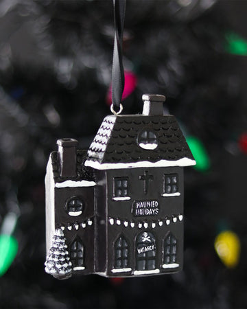 Close-up of the Rebel Romance Haunted Holiday House resin Christmas decoration hanging from a black ribbon on a dark Christmas tree, with snow-covered roof details, twinkly lights, and gothic “Haunted Holidays” and “VACANCY” signs.