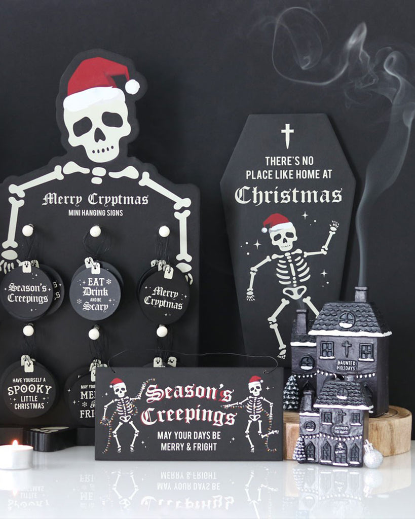 A gothic Christmas flat lay featuring black skeleton-themed decorations from Rebel Romance, including a mini haunted house ornament, coffin sign saying “There’s No Place Like Home at Christmas,” and dark festive signs with messages like “Season’s Creepings” and “Merry Cryptmas.”