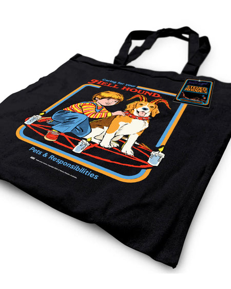 A black tote bag featuring Steven Rhodes’ “Caring for Your Hell Hound” design. The artwork showcases a boy kneeling next to a beagle with red horns, sitting in the middle of a summoning circle surrounded by lit candles. The text reads “Caring for Your Hell Hound” and “Pets & Responsibilities.”