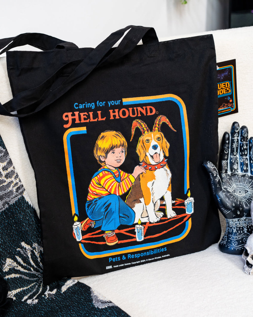 A black tote bag featuring Steven Rhodes’ “Caring for Your Hell Hound” design. The artwork showcases a boy kneeling next to a beagle with red horns, sitting in the middle of a summoning circle surrounded by lit candles. The text reads “Caring for Your Hell Hound” and “Pets & Responsibilities.”
