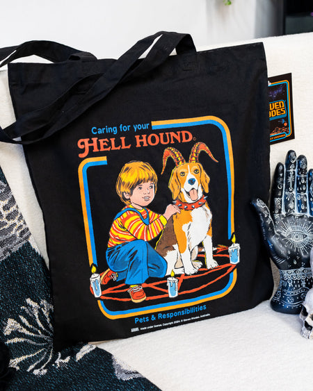 A black tote bag featuring Steven Rhodes’ “Caring for Your Hell Hound” design. The artwork showcases a boy kneeling next to a beagle with red horns, sitting in the middle of a summoning circle surrounded by lit candles. The text reads “Caring for Your Hell Hound” and “Pets & Responsibilities.”