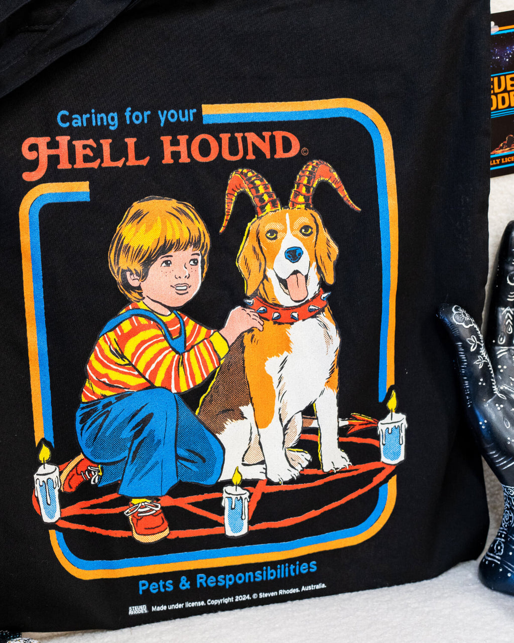 A black tote bag featuring Steven Rhodes’ “Caring for Your Hell Hound” design. The artwork showcases a boy kneeling next to a beagle with red horns, sitting in the middle of a summoning circle surrounded by lit candles. The text reads “Caring for Your Hell Hound” and “Pets & Responsibilities.”