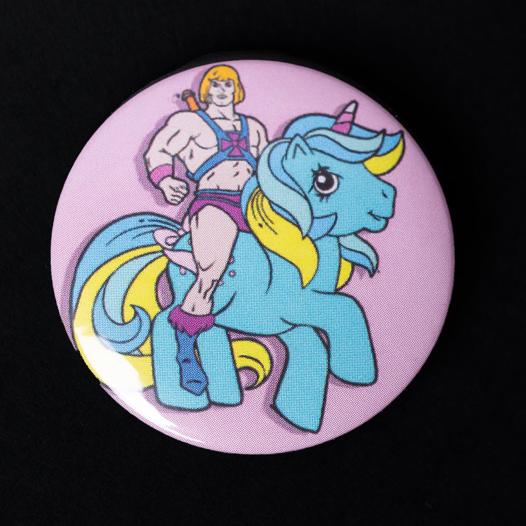 A 38mm button badge featuring He-Man riding a blue unicorn with a rainbow mane and tail on a pastel pink background. The high-gloss finish highlights the playful and nostalgic pop culture design. Created by Bite Your Granny, this pin is perfect for adding a quirky touch to any accessory.