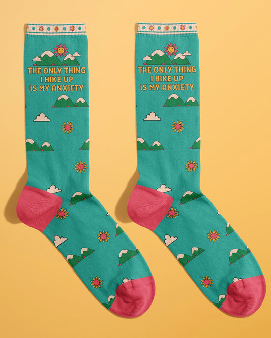Flat lay of a pair of teal novelty socks from Rebel Romance with pink toes and heels. The socks are covered in cartoon clouds, flowers, and mountains with the slogan "The only thing I hike up is my anxiety." Designed by FUN CLUB for a fun, quirky look.