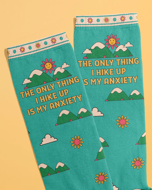 Close-up of teal crew socks by Rebel Romance featuring the phrase "The only thing I hike up is my anxiety" in bold yellow text, with cartoon mountains, clouds, and a smiling pink daisy sun. The ribbed trim is decorated with cheerful flowers. Designed by FUN CLUB.