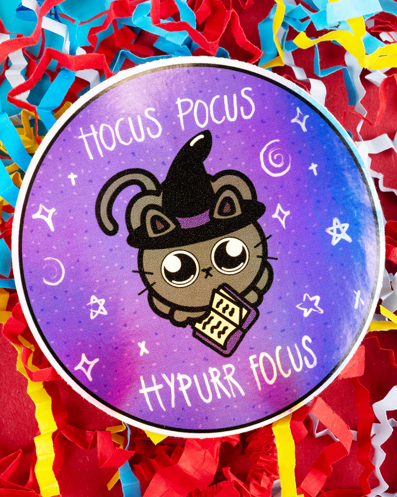 Whimsical circular sticker with a holographic finish, showing a black witch cat holding an open book under the text “Hocus Pocus Hypurr Focus.” Surrounded by stars and magical symbols on a purple background. A quirky gift idea from Rebel Romance and Innabox, great for neurodivergent folks or fans of alternative style and magic.