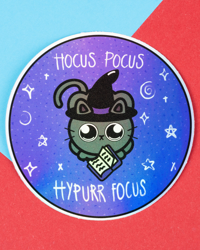 Round holographic vinyl sticker featuring a cute cartoon black cat wearing a witch hat and holding a spellbook. The sticker has a purple gradient background with stars and reads “Hocus Pocus Hypurr Focus.” Perfect for celebrating hyperfocus and neurodivergent pride. Product by Innabox, sold by Rebel Romance, a UK brand for alternative fashion and gifts.