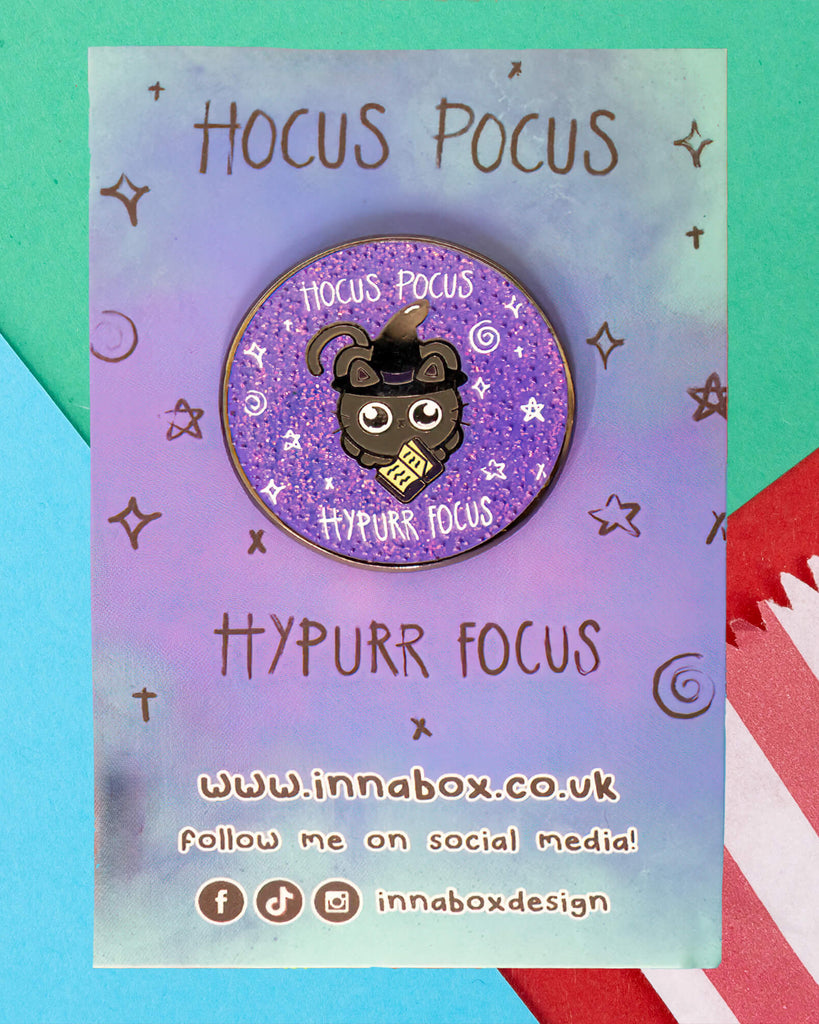 A purple glitter enamel pin by Innabox, sold by Rebel Romance, featuring a cute black cat in a witch hat holding a book. Surrounded by stars and sparkles, the pin reads “Hocus Pocus Hypurr Focus.” Displayed on colourful branded backing with “Hocus Pocus Hypurr Focus” and innabox.co.uk details.