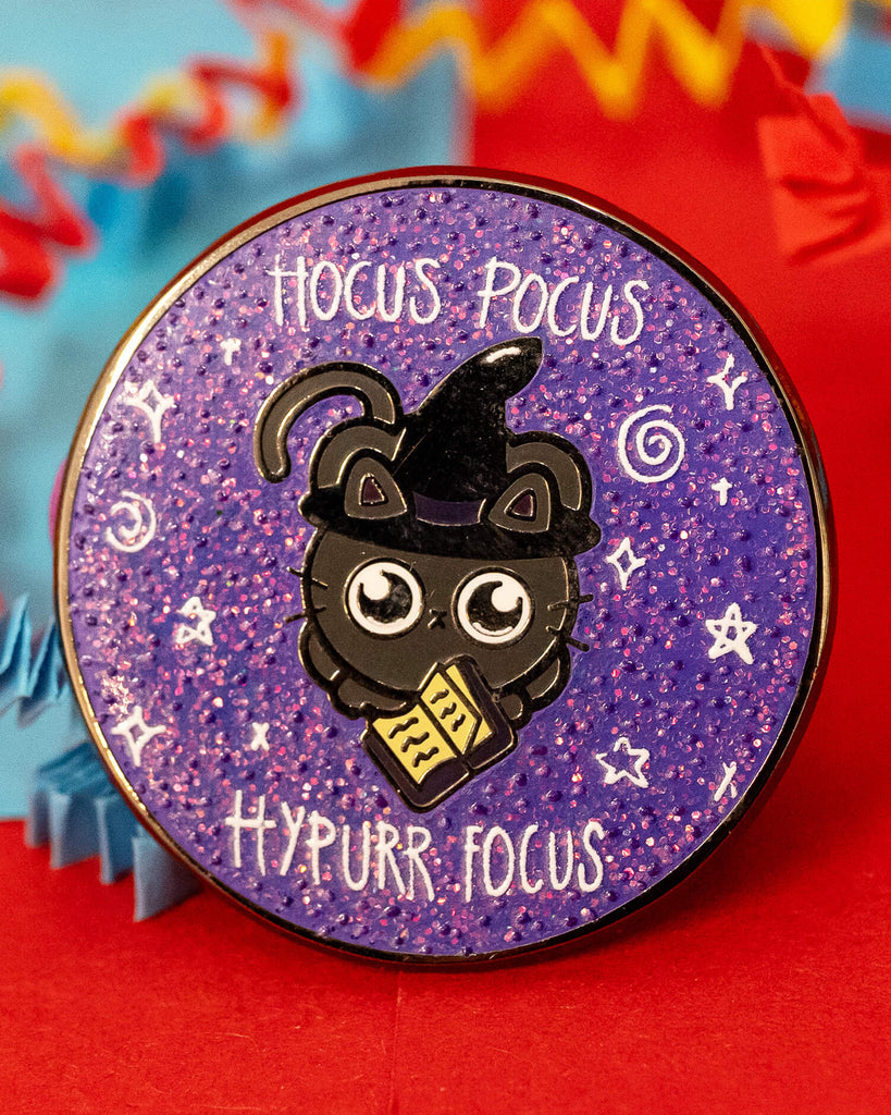 Close-up of a purple glitter enamel pin from Rebel Romance by Innabox. The design shows a black cat in a witch hat holding a spellbook, with the words “Hocus Pocus Hypurr Focus” in white above and below. Surrounded by hand-drawn stars and sparkles on a textured glitter background.