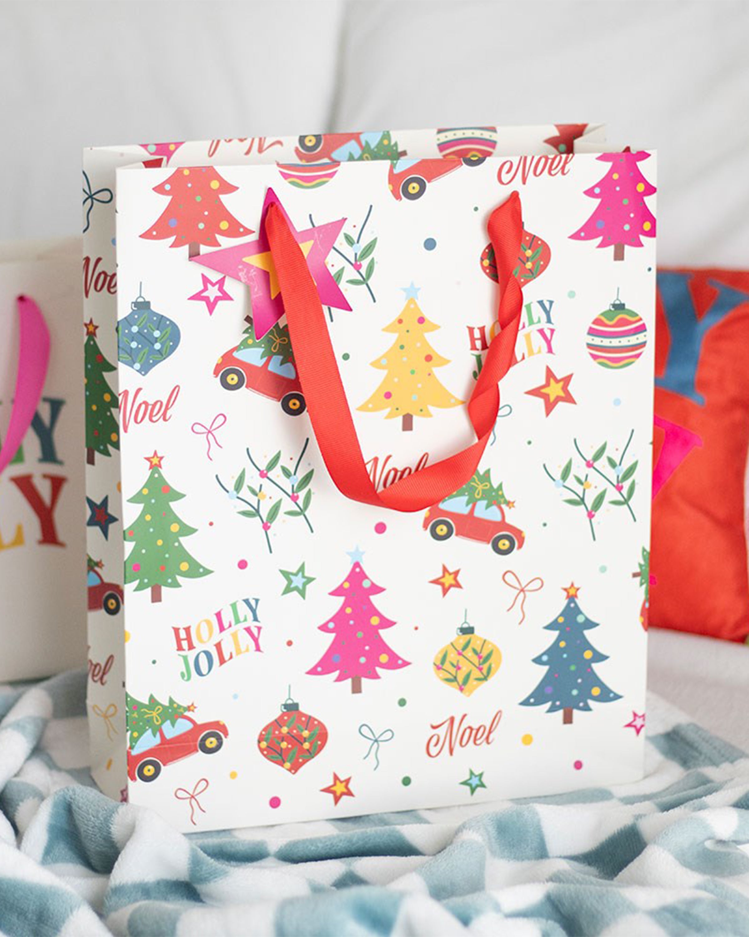 A Rebel Romance Nostalgic Christmas Print Gift Bag styled on a cosy blanket with vibrant festive patterns, red ribbon handles, and fun retro Christmas illustrations, perfect for wrapping presents in a playful, alternative style.