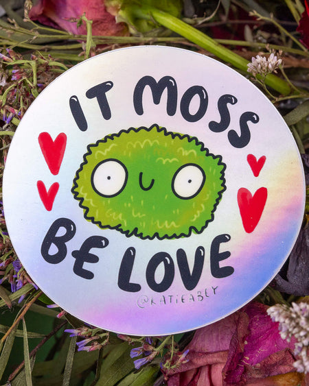 A round holographic vinyl sticker by Katie Abey, sold by Rebel Romance. The design features a cute green moss character with big eyes and a smile, surrounded by red cartoon hearts. Text above and below reads: “It Moss Be Love.” The rainbow shimmer of the holographic background catches the light, making this quirky sticker shine with charm and positive vibes.