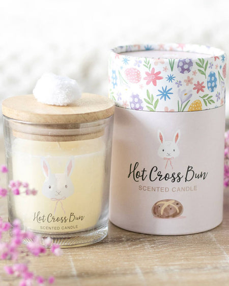 A cosy lifestyle shot of the candle and its matching gift box placed on a wooden surface with delicate pink flowers in the background. The candle’s pom-pom lid and the box’s cheerful springtime design are clearly visible.