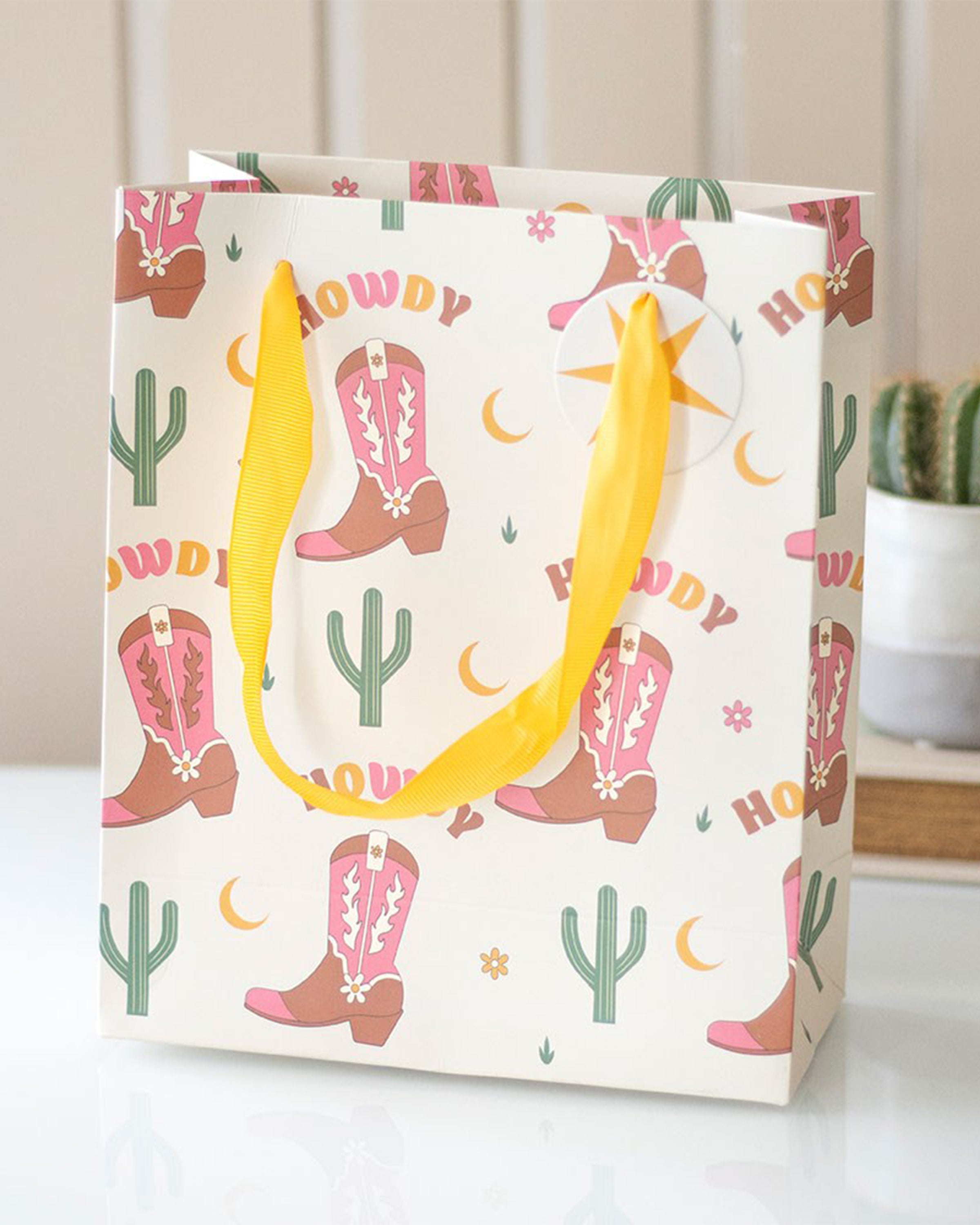 Rebel Romance medium “Howdy” gift bag styled on a table, with bold yellow handles, pink cowboy boots, cacti and moon print, perfect for Western‑themed gifting.