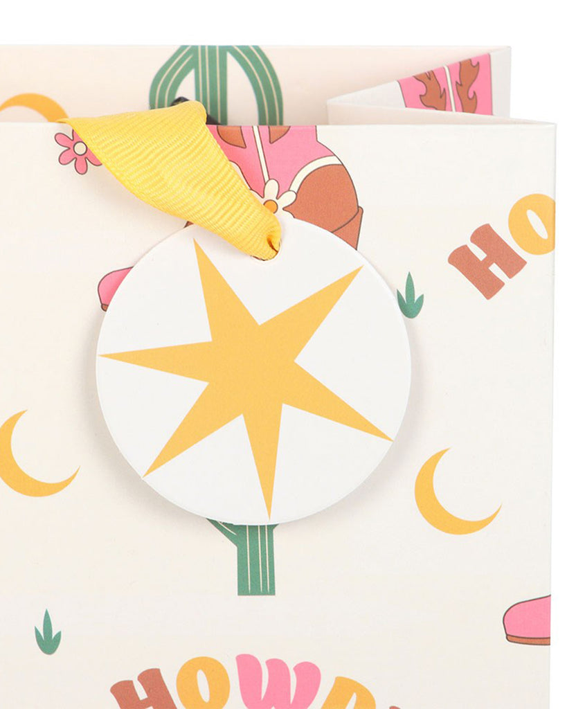 Close‑up of Rebel Romance’s medium cowboy boot gift bag, showing a yellow ribbon handle and round star tag against a cream background with “Howdy” text and cacti print.