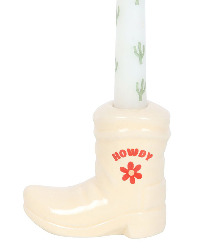 Close-up of the white cowboy boot candle holder, featuring the word "Howdy" in bold red letters with a retro red flower below. The cactus-patterned taper candle fits snugly into the boot. Sold by Rebel Romance, embracing extraordinary style and Western flair.