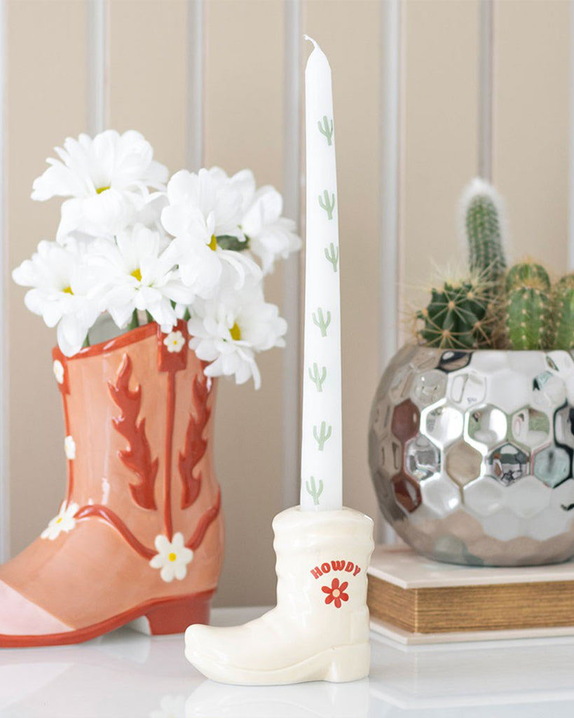 Styled shot of the Rebel Romance cowboy boot candle holder on a white shelf. It’s paired with a pink boot vase filled with daisies and a silver geometric planter holding cacti, highlighting the holder’s Western charm and playful aesthetic.
