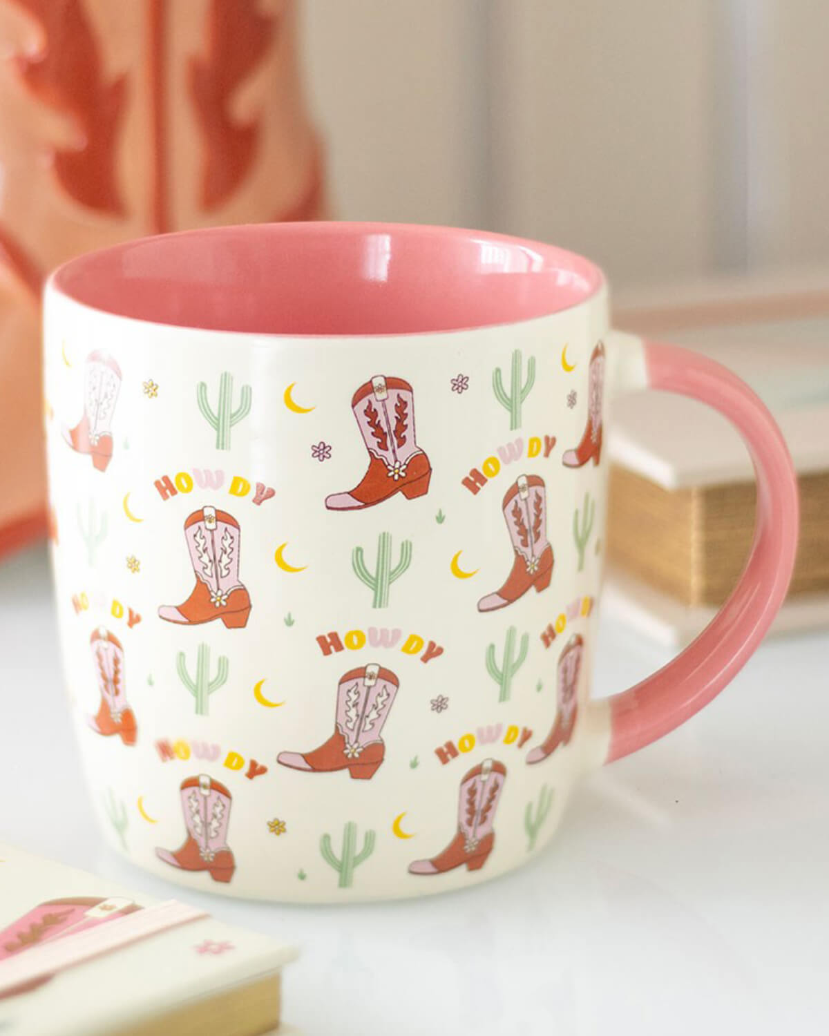 Image 3 (Lifestyle Shot) – The cowboy boot print mug is placed on a table with Western-themed decor, including a cowboy boot vase and books.