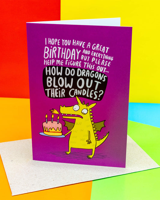 A bright purple Rebel Romance A6 greeting card designed by Katie Abey, featuring a funny yellow dragon holding a pink birthday cake with lit candles. The text reads “How do dragons blow out their candles?” A quirky, alternative birthday card perfect for millennials who love unique designs.