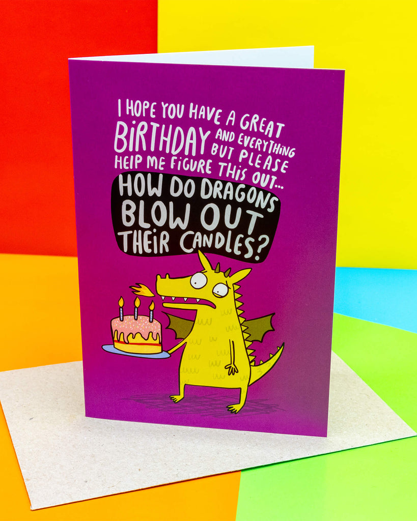 A bright purple Rebel Romance A6 greeting card designed by Katie Abey, featuring a funny yellow dragon holding a pink birthday cake with lit candles. The text reads “How do dragons blow out their candles?” A quirky, alternative birthday card perfect for millennials who love unique designs.