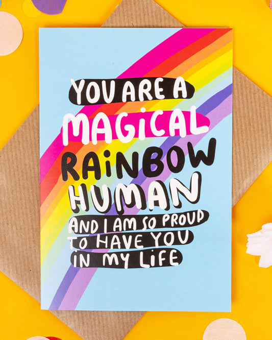 A colourful A6 greeting card from Rebel Romance designed by Katie Abey, featuring bold rainbow stripes and playful typography that reads “You are a magical rainbow human and I am so proud to have you in my life.” Set on a light blue background, the card is shown with a kraft envelope and confetti on a yellow backdrop. A joyful and inclusive design perfect for gifting.