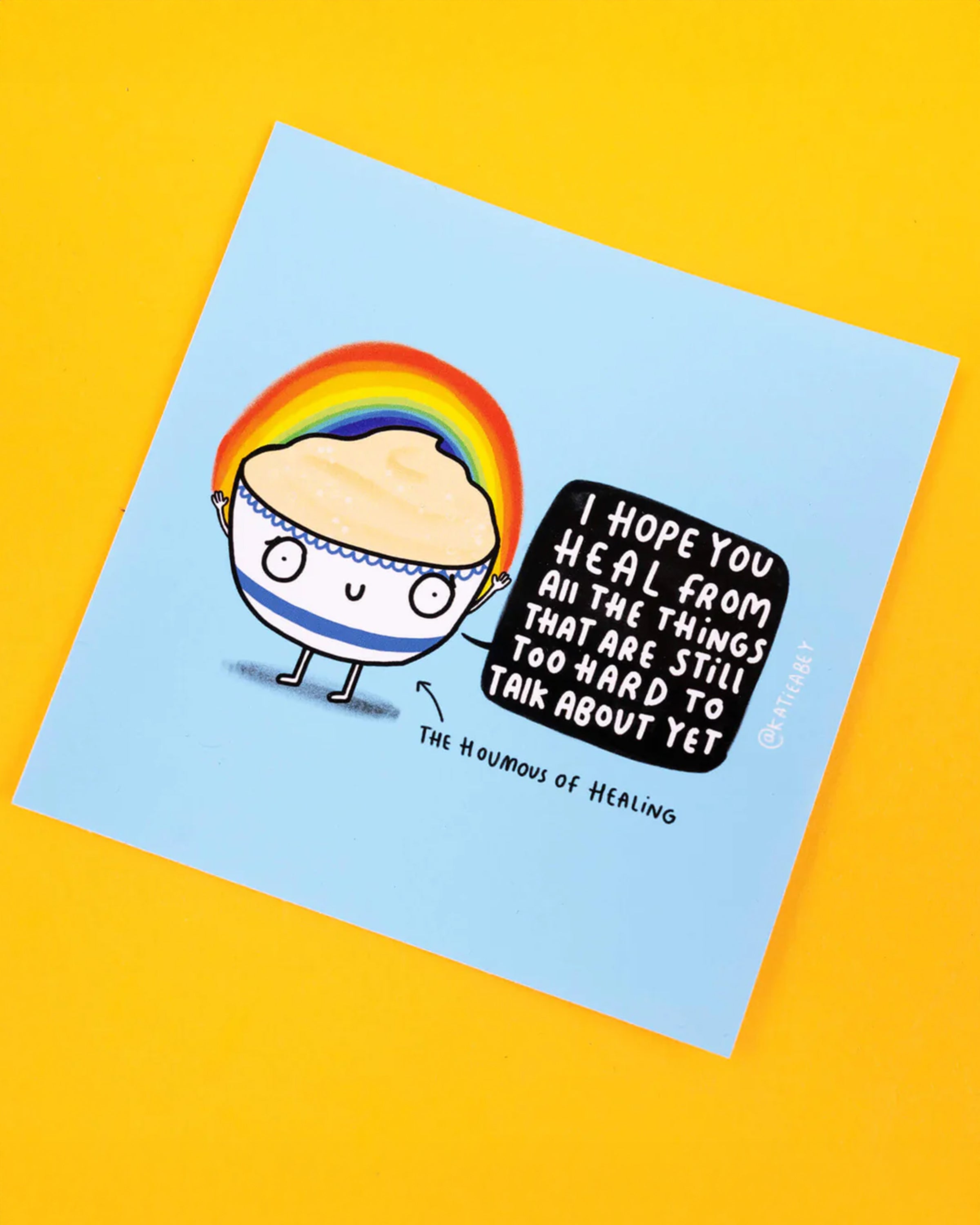 Katie Abey vinyl sticker featuring a cute, cartoon hummus character, with a rainbow arching above its head. The character holds a speech bubble that says, 'I Hope You Heal From All The Things That Are Still Too Hard To Talk About Yet', with the label 'The Houmous of Healing' beneath.