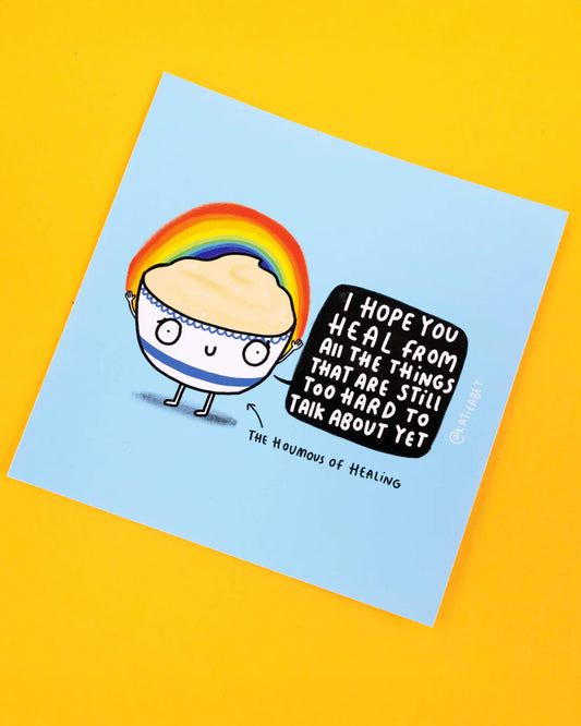 Katie Abey vinyl sticker featuring a cute, cartoon hummus character, with a rainbow arching above its head. The character holds a speech bubble that says, 'I Hope You Heal From All The Things That Are Still Too Hard To Talk About Yet', with the label 'The Houmous of Healing' beneath.