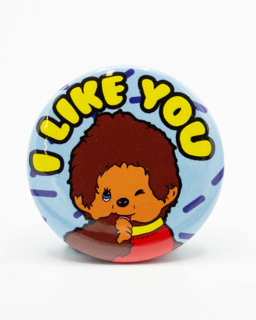 A circular button pin badge photographed against a white background. The badge shows a cute Chick-A-Boo inspired character winking and sucking its thumb beneath bright yellow bubble text that reads “I LIKE YOU.” A fun, retro design by Bite Your Granny, available from Rebel Romance – a small UK business celebrating alternative aesthetics and quirky gifts.