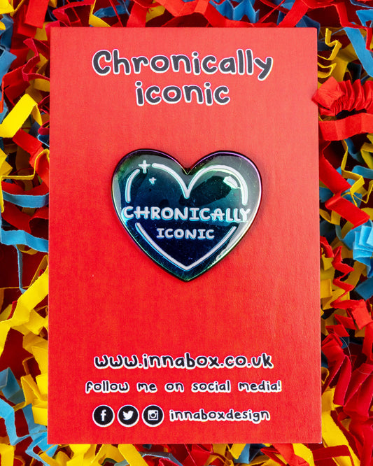 A rainbow heart-shaped enamel pin that reads “Chronically Iconic” in bold white lettering, displayed on a red card with colourful confetti background. The pin is designed by Innabox and sold by Rebel Romance, a small UK business celebrating alternative fashion, inclusivity, and millennial style.