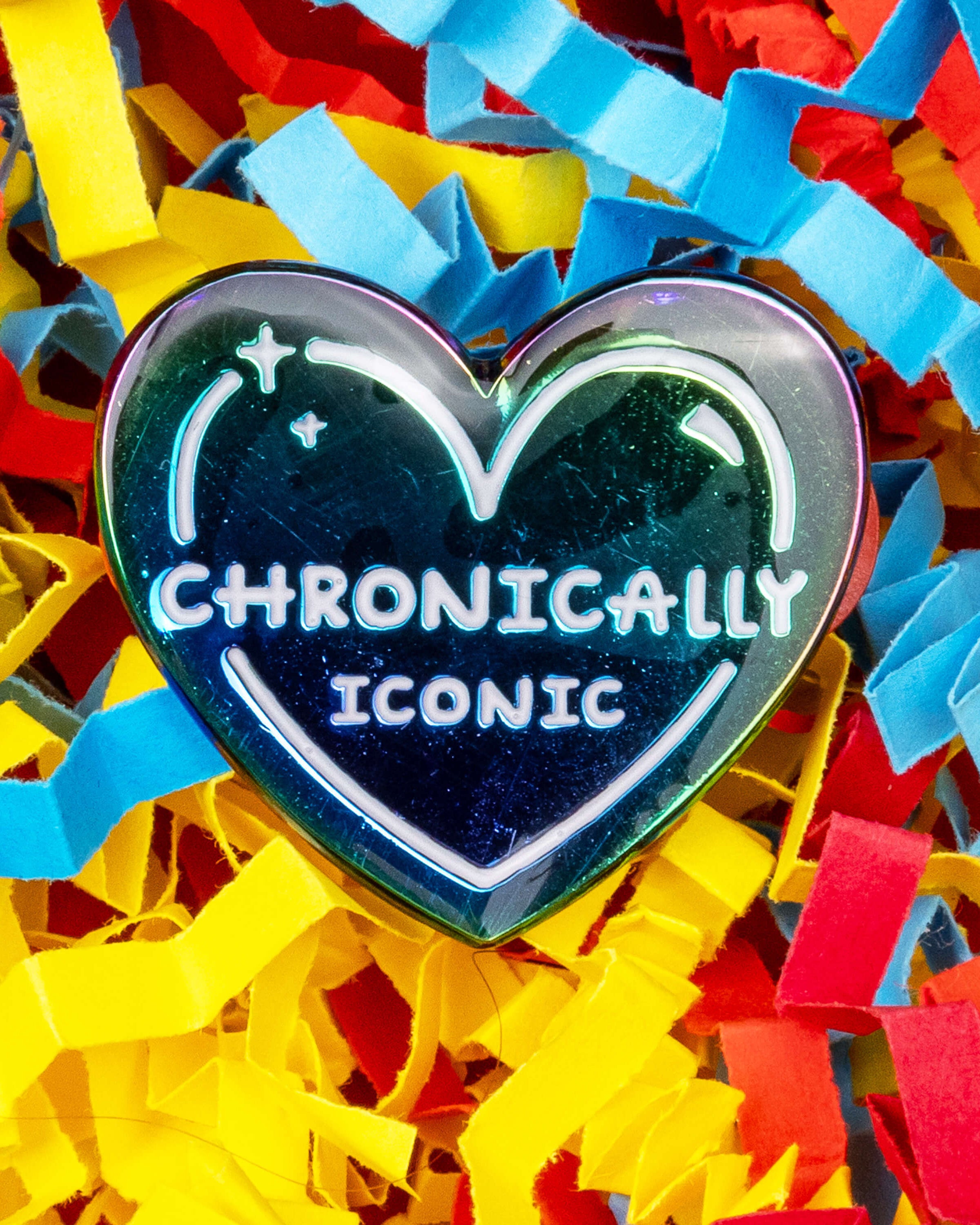 Close-up of the “Chronically Iconic” rainbow heart enamel pin, featuring colourful shiny metal edging and white lettering. The pin rests on a red, blue, and yellow crinkle confetti paper background. Sold by Rebel Romance, a UK brand celebrating individuality and alternative aesthetics.