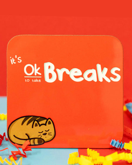 Close-up of the It's Ok To Take Breaks Cat Coaster by Innabox. The square coaster has rounded corners and a bright red background. The design features white, playful text reading "It's Ok to take Breaks," with a cute illustration of a curled-up, sleeping brown tabby cat in the bottom left corner. Colourful red, blue, and yellow crepe paper surrounds the coaster. Available on Rebel Romance 