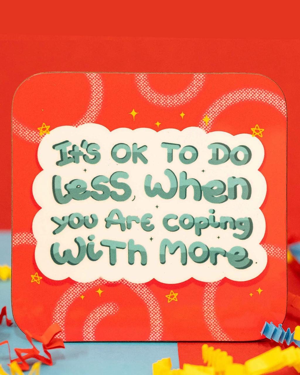 Close-up of the It's Ok To Do Less Coaster by Innabox. The square coaster has rounded corners and a bold red background with swirly patterns, tiny stars, and a small rainbow in the top right. The design features a fluffy white speech bubble with green text reading, 