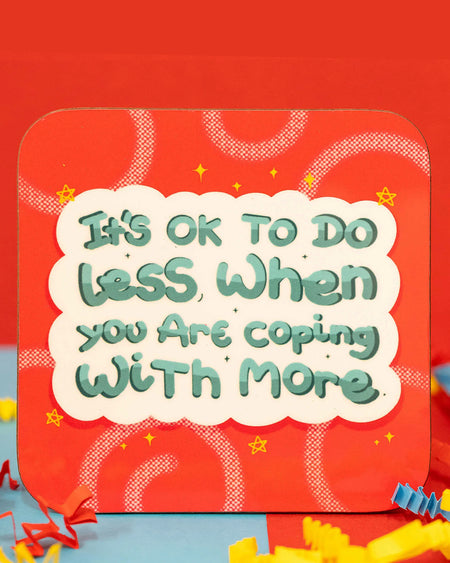 Close-up of the It's Ok To Do Less Coaster by Innabox. The square coaster has rounded corners and a bold red background with swirly patterns, tiny stars, and a small rainbow in the top right. The design features a fluffy white speech bubble with green text reading, "It's OK to do less when you are coping with more." Colourful red, blue, and yellow crepe paper surrounds the coaster. Available on Rebel Romance